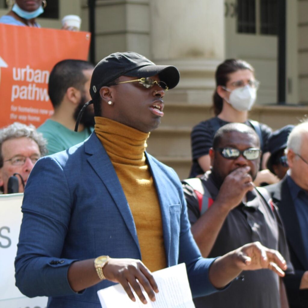 A man in a blue blazer and yellow turtleneck speaks at a press conference on the steps of a building. Behind him, supporters stand in solidarity, holding signs for "Urban Pathways" and "Neighbors Together." conference. Behind him, supporters stand on the steps of a building, holding signs for "Urban Pathways" and "Neighbors Together."