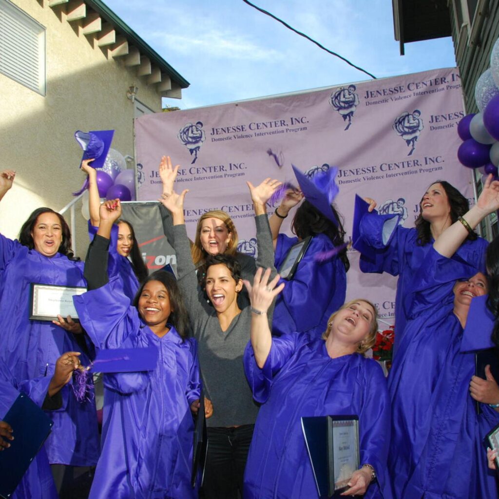 A group of women in purple graduation gowns joyfully celebrate outdoors, holding certificates and throwing their caps in the air. They stand in front of a pink "Jenesse Center, Inc." banner and purple balloons.