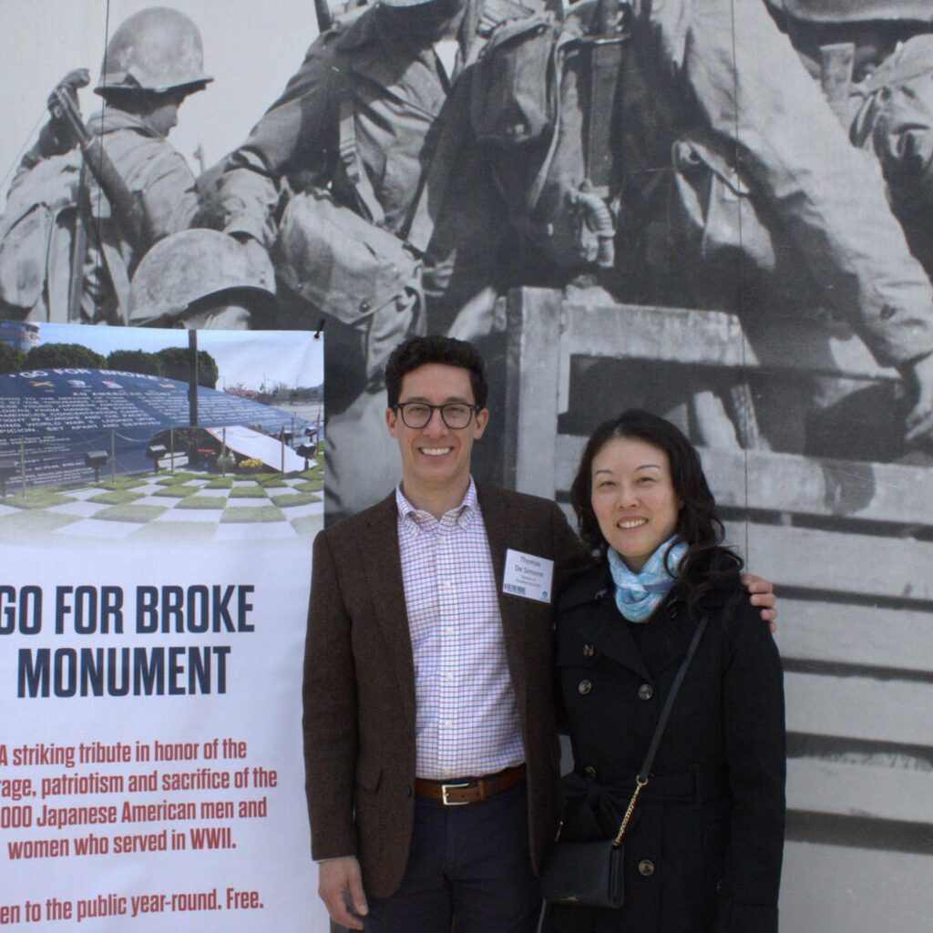 A man and woman smile for a photo, standing next to a large sign for the "Go For Broke Monument." The sign honors the 33,000 Japanese American men and women who served in WWII, and behind them is a black-and-white mural of soldiers.