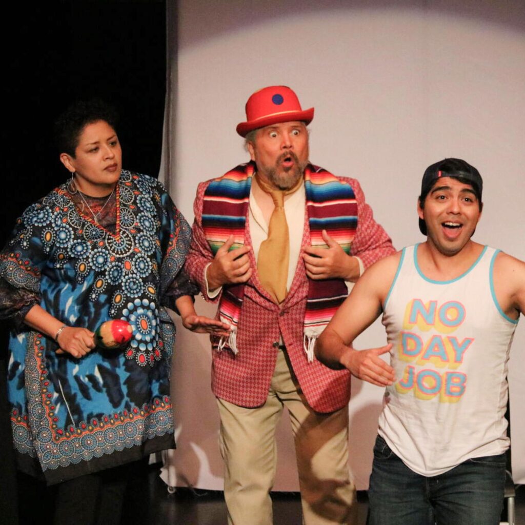 Three actors with animated expressions perform on a stage in colorful costumes. One man wears a red hat and patterned scarf, while another wears a tank top that reads, "NO DAY JOB."