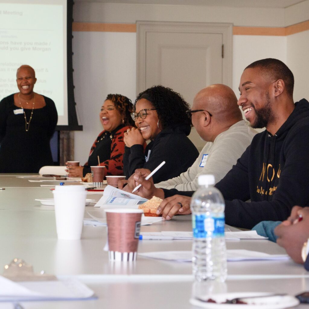 A group of adults seated along a conference table laugh and smile during a presentation. A woman stands smiling at the front of the room next to a projector screen.