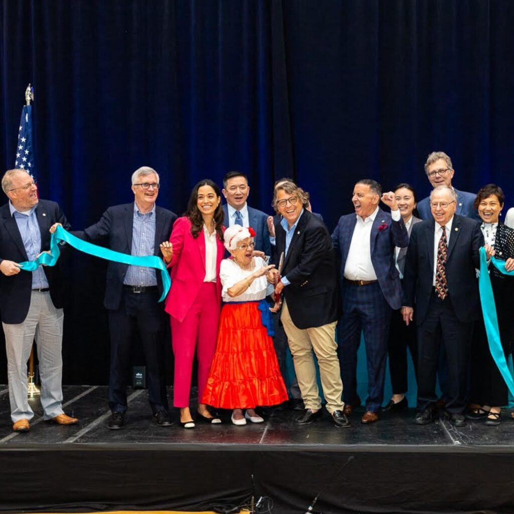 A large group of people smiles and celebrates on a stage during a ribbon-cutting ceremony. An older woman in a festive red skirt and white top is at the center, holding scissors to cut a light blue ribbon held by others in the group.