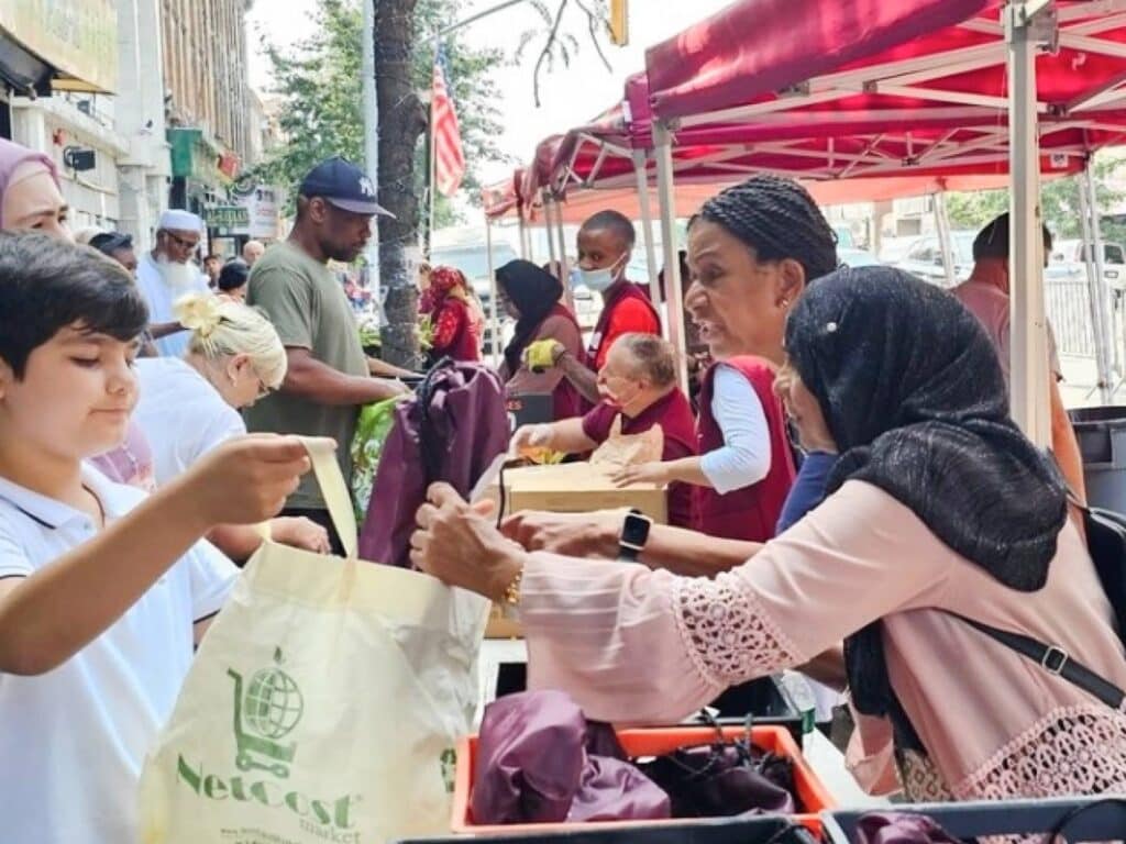 Volunteers at an outdoor "School Supplies Giveaway" hand out purple drawstring bags to a diverse group of community members from under a red tent on a city street.