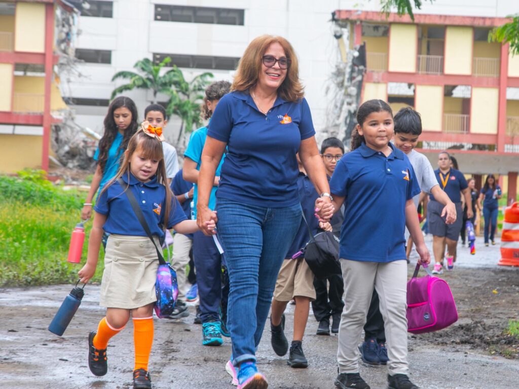 A smiling woman in a blue polo shirt and jeans holds hands with two young schoolgirls, leading a larger group of children and adults. They walk together on a path with large, partially damaged school buildings in the background.