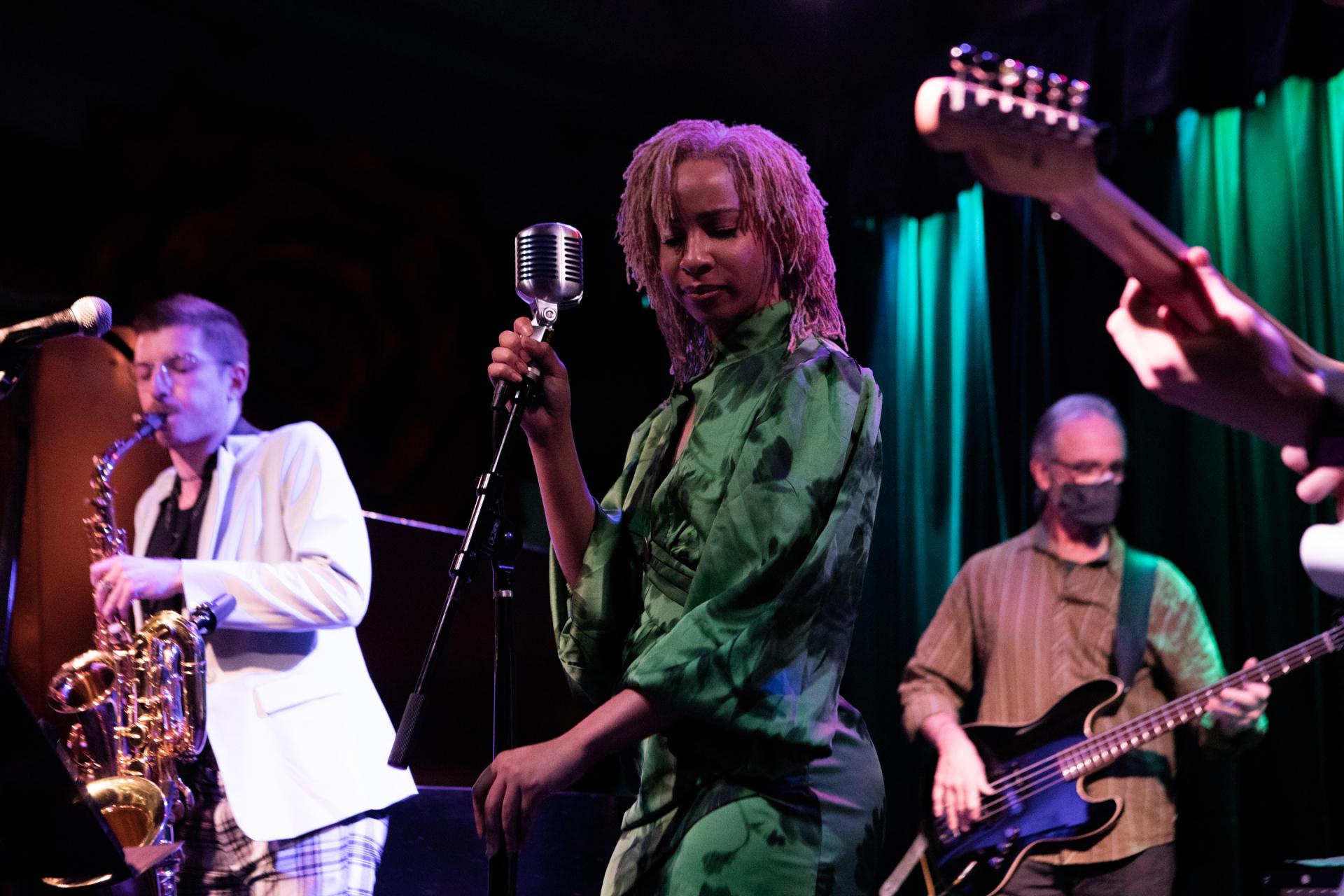 A female singer with pink-tinted locs holds a vintage microphone while performing on a dimly lit stage. Her band, including a saxophonist and a bassist, plays behind her.