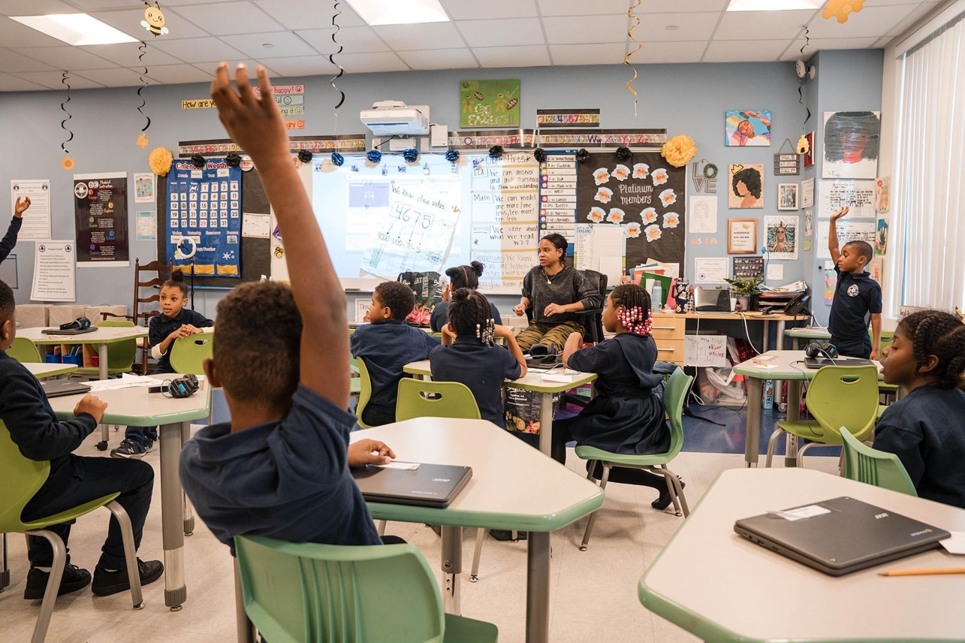 In a decorated elementary classroom, a teacher leads a lesson as students sit at their desks. Several students have their hands raised to answer, including a boy in the foreground whose arm is stretched high.