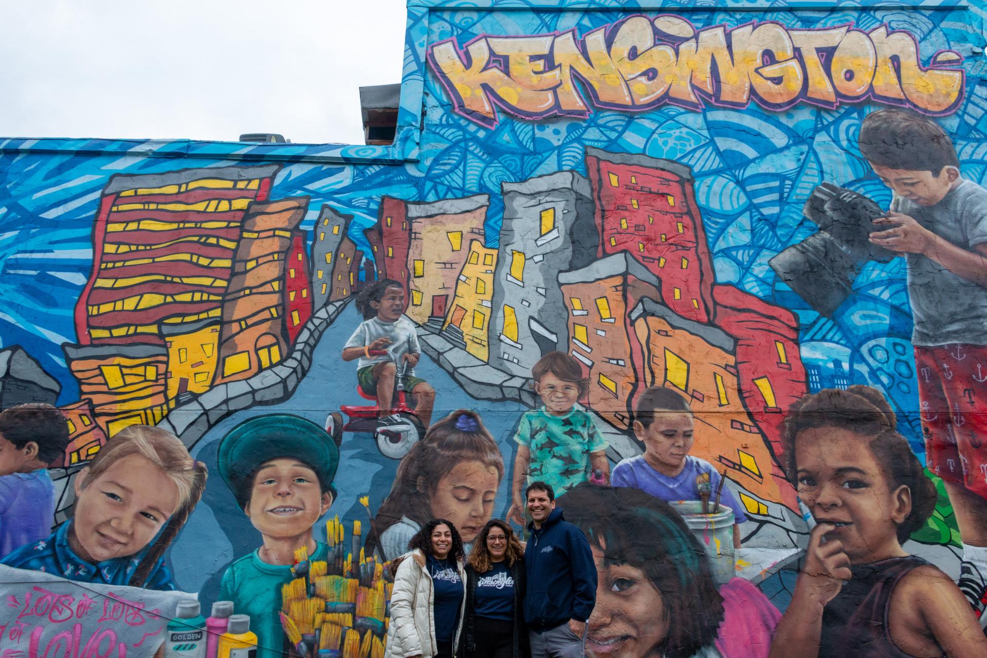 Three smiling adults stand together for a photo in front of a large, colorful mural that reads "Kensington" and depicts portraits of children and stylized city buildings.