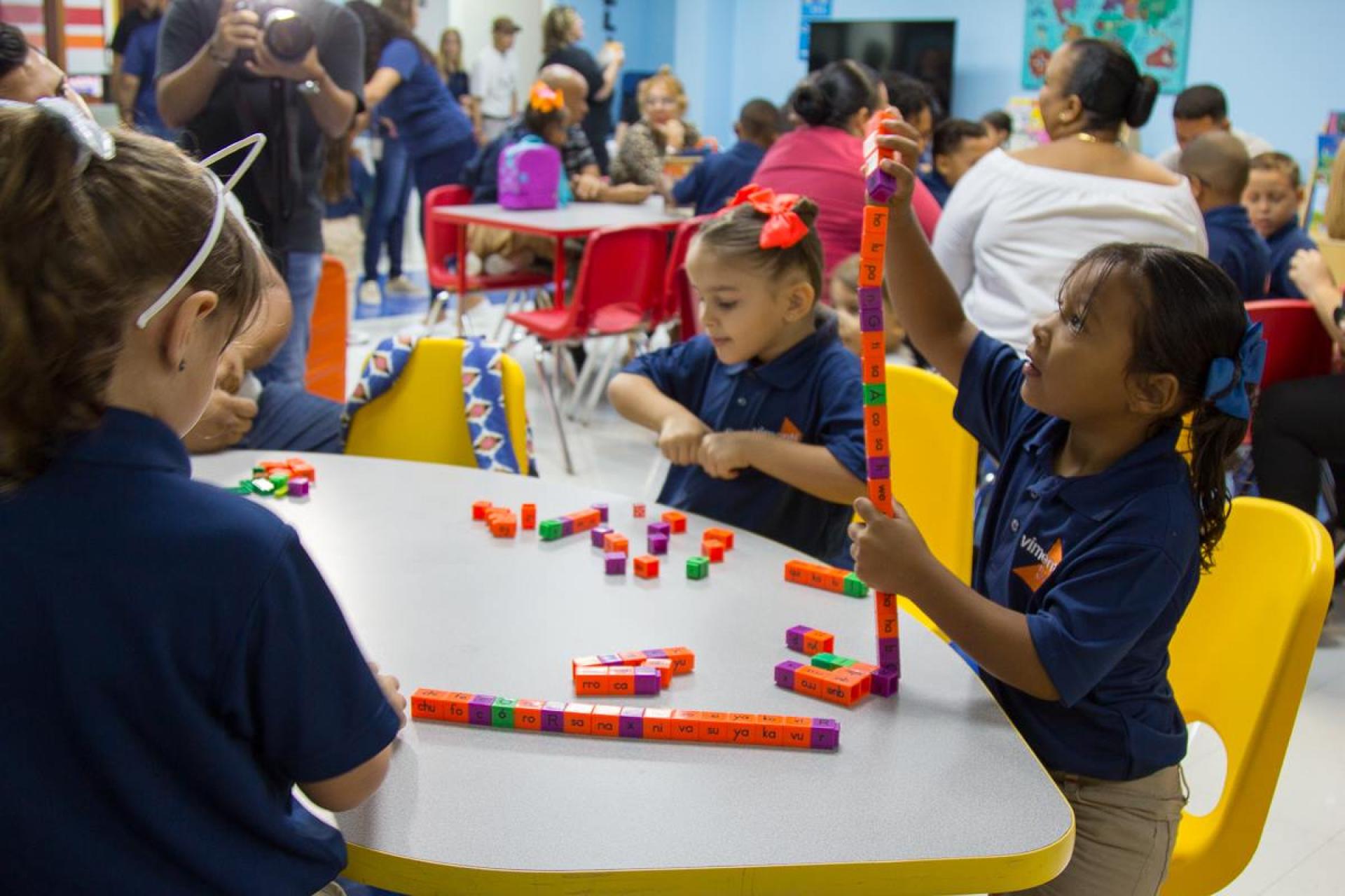 Young girls in blue school uniforms sit around a table in a busy classroom, playing with colorful, interlocking learning blocks. One girl proudly holds up a tall tower she built from the blocks, while her classmates are also engaged with the activity.