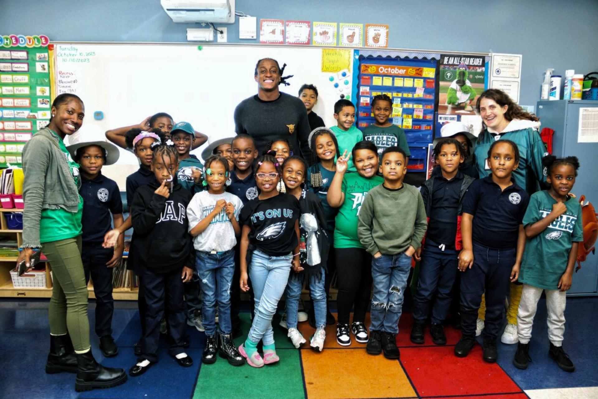 A large group of elementary school children and three adults smile and pose for a group photo inside a colorful classroom. Many of the children and adults are wearing Philadelphia Eagles fan apparel.