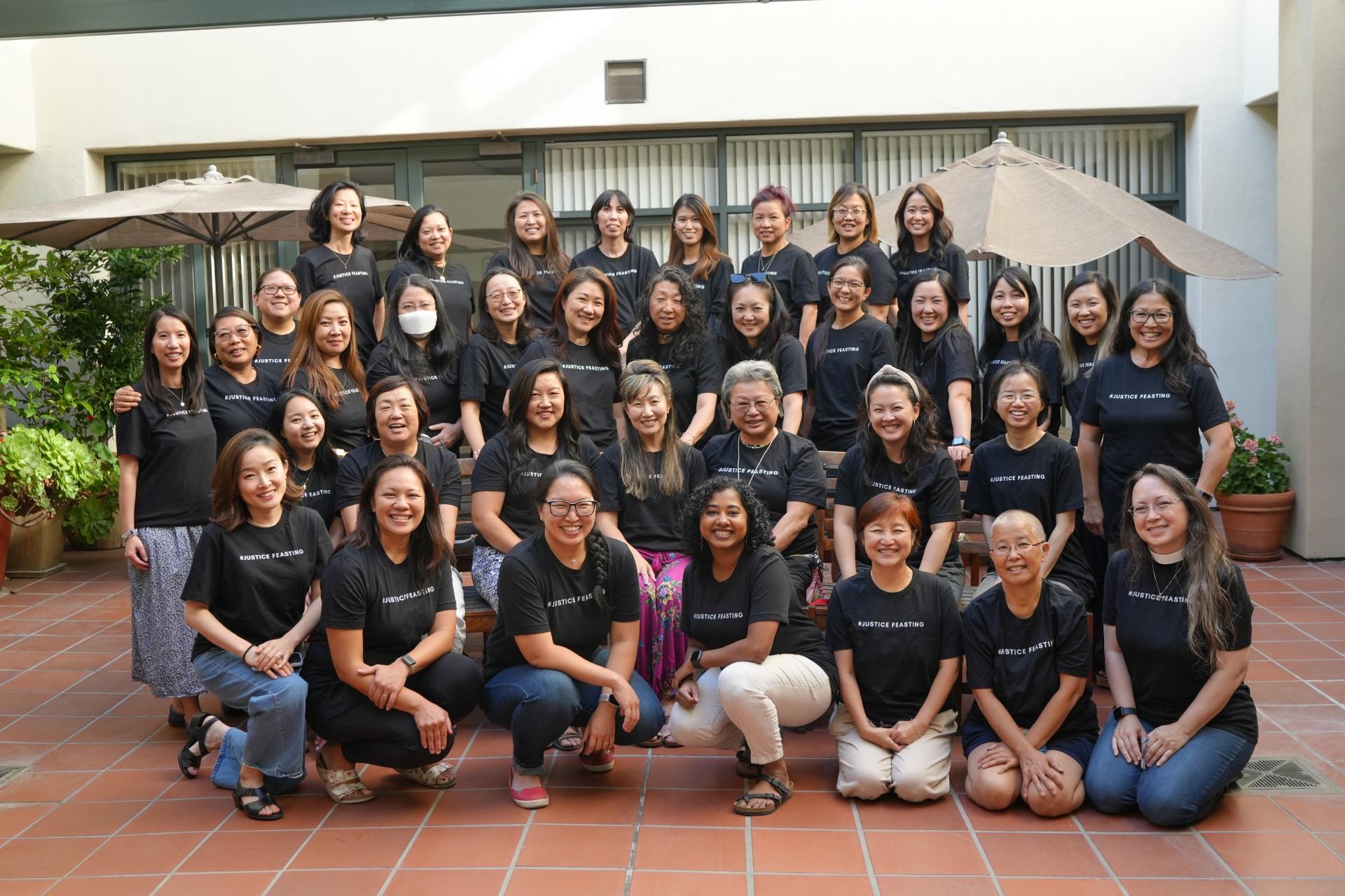 A large group of women, all wearing matching black t-shirts, pose for a group photo in a tiled outdoor courtyard. They are arranged in several rows, smiling for the camera.
