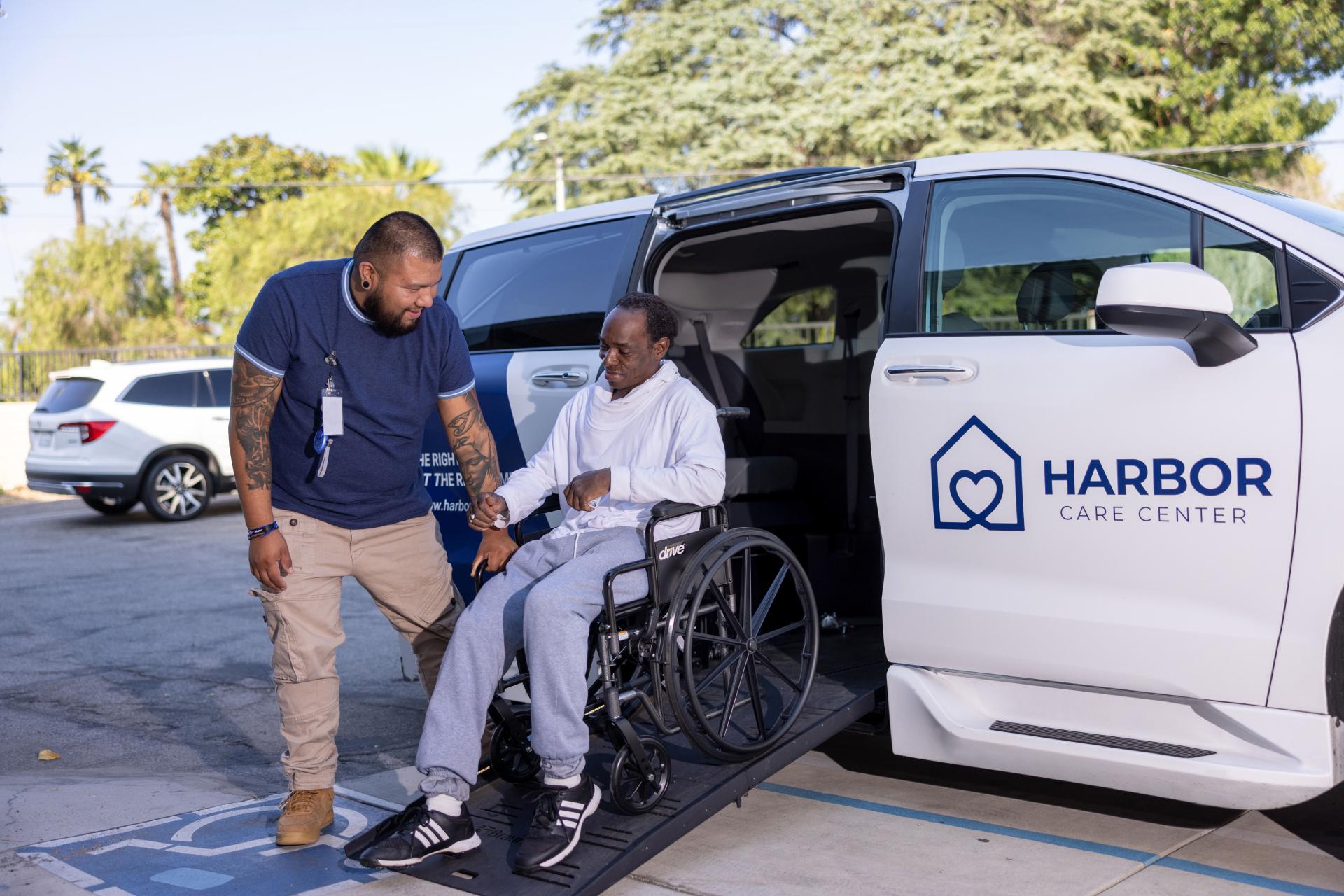 A caregiver from Harbor Care Center smiles as he assists a person in a wheelchair up the ramp into an accessible white transport van.
