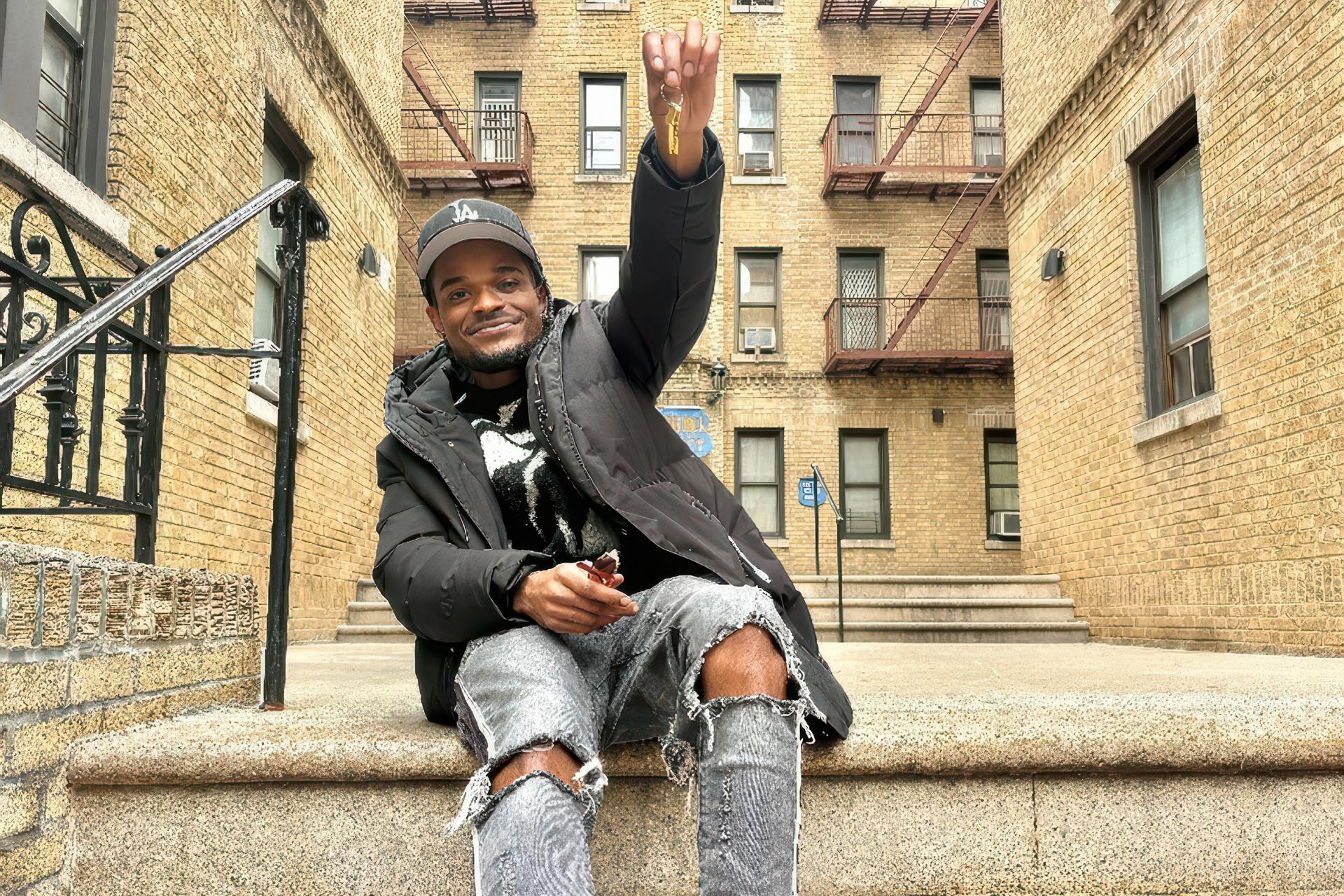 A smiling young man sits on the front steps of a large brick apartment building and proudly holds up a set of keys.