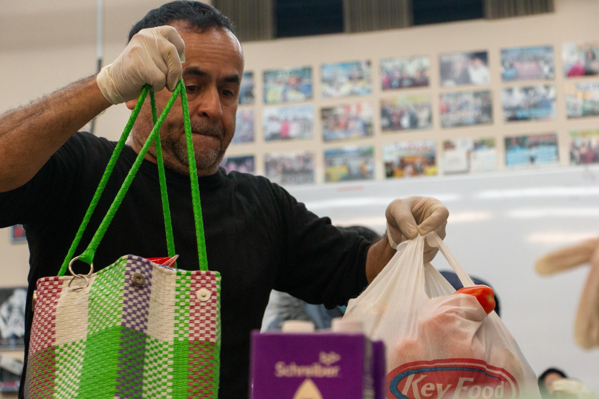 A man wearing disposable gloves handles groceries at a food bank, holding a colorful woven tote and a plastic grocery bag filled with food.