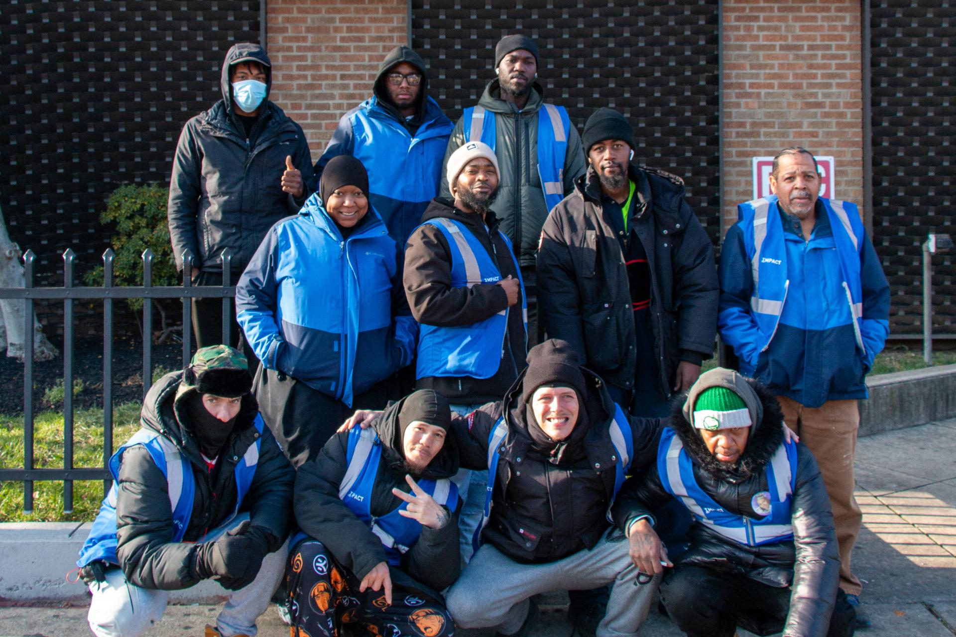 A group of about ten workers dressed in warm jackets and blue vests pose for a photo on the sidewalk. They are standing and crouching in front of a brick building.
