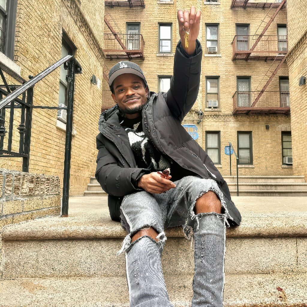 A smiling young man sits on the front steps of a large brick apartment building and proudly holds up a set of keys.