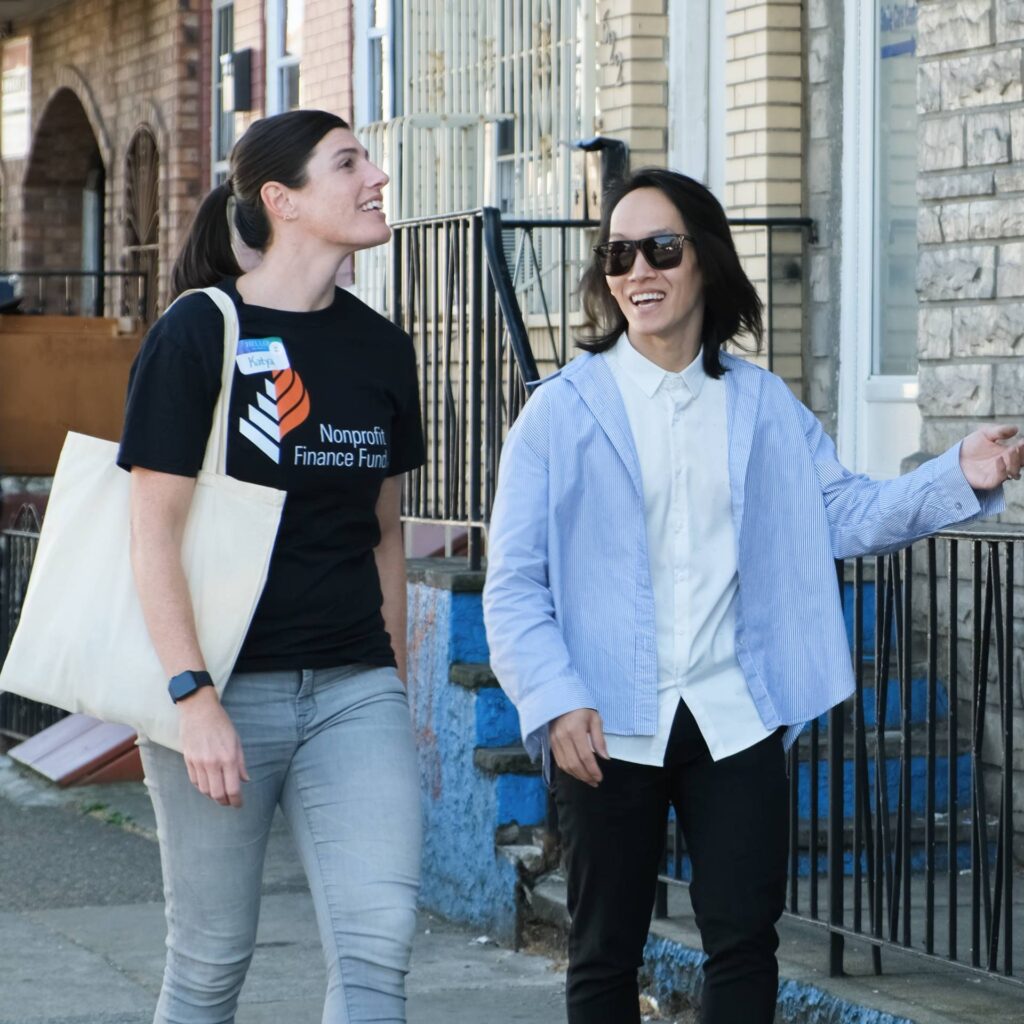 A woman in a "Nonprofit Finance Fund" t-shirt and a person with long hair and sunglasses walk together down a city sidewalk. They are smiling and engaged in conversation as they pass by row houses.