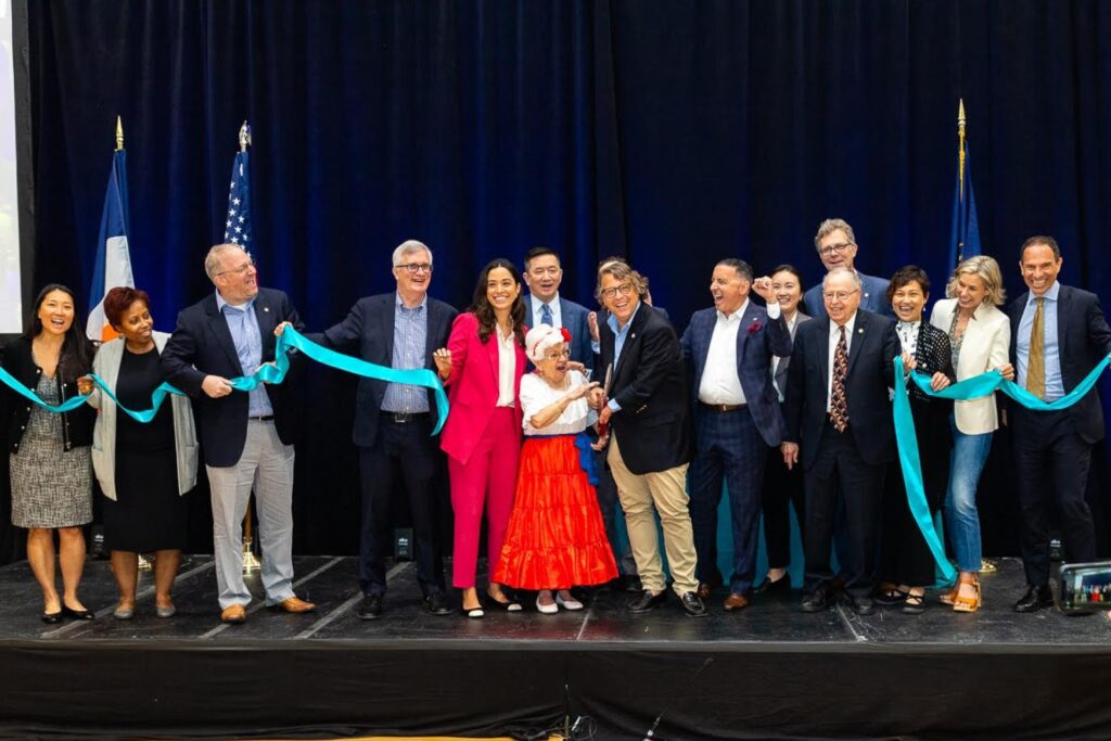 A large group of people smiles and celebrates on a stage during a ribbon-cutting ceremony. An older woman in a festive red skirt and white top is at the center, holding scissors to cut a light blue ribbon held by others in the group.