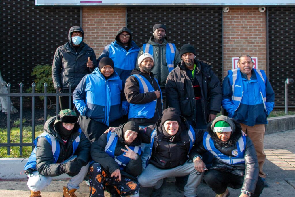 A group of about ten workers dressed in warm jackets and blue vests pose for a photo on the sidewalk. They are standing and crouching in front of a brick building.