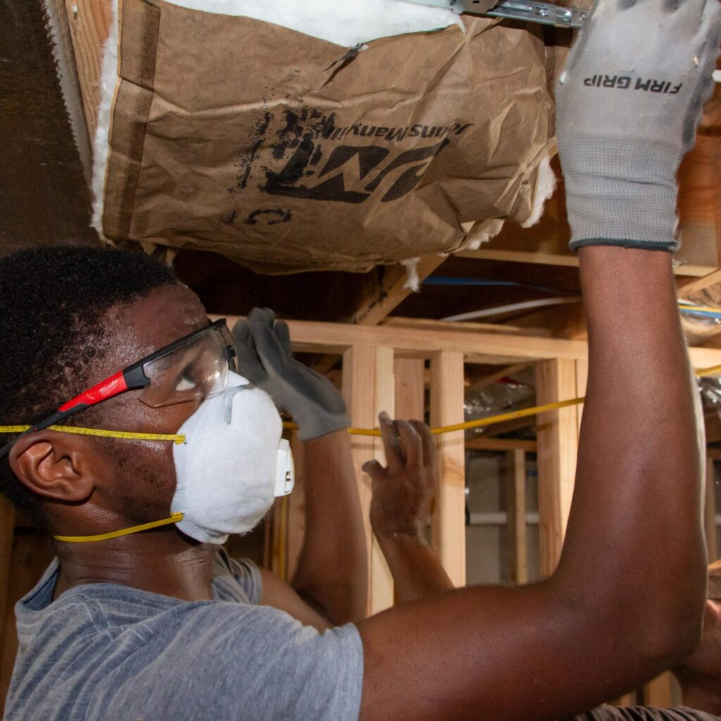 A person wearing safety goggles, a mask, and gloves installs insulation in the ceiling of a room under construction. Another worker is visible in the background.