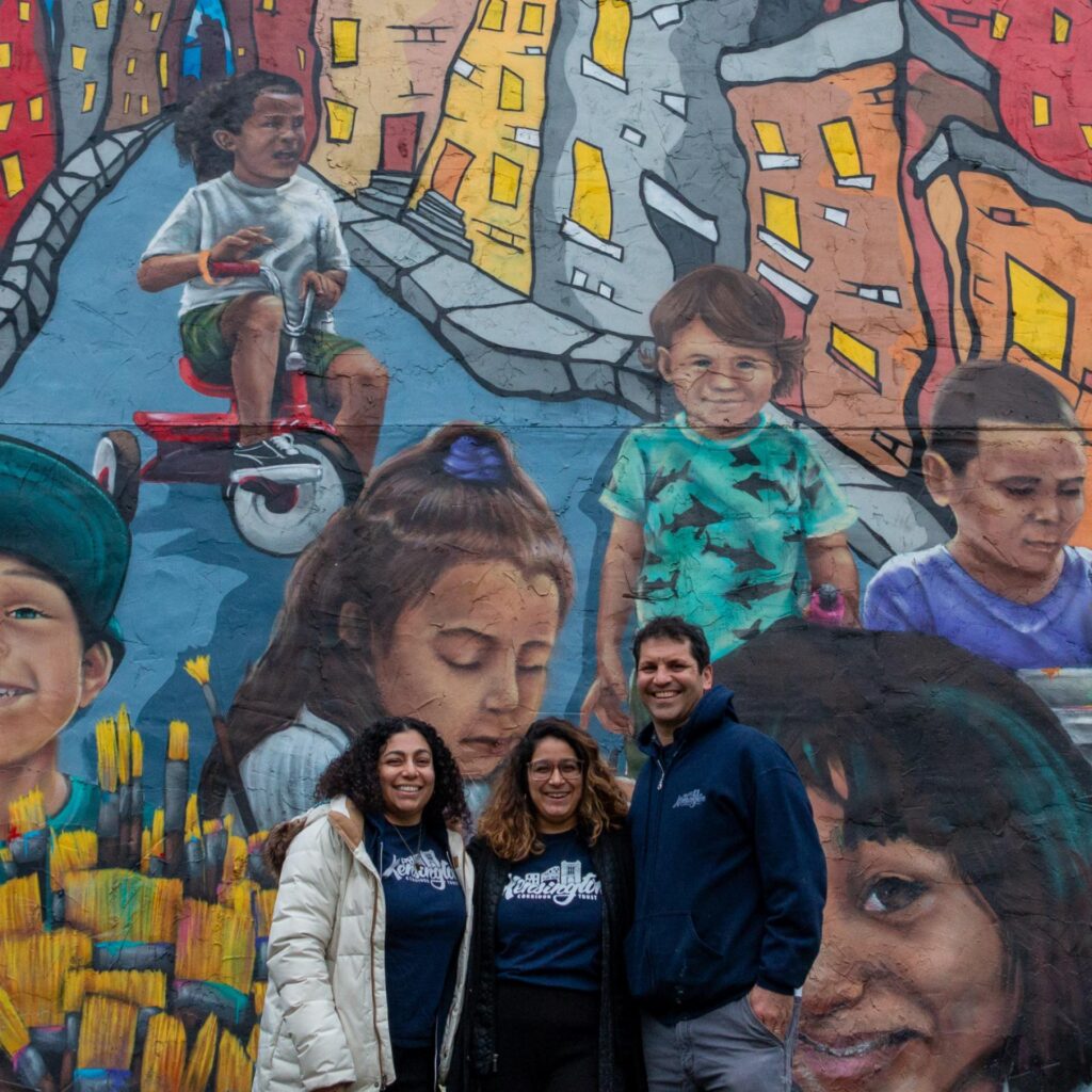 Three smiling adults stand together for a photo in front of a large, colorful mural that reads "Kensington" and depicts portraits of children and stylized city buildings.