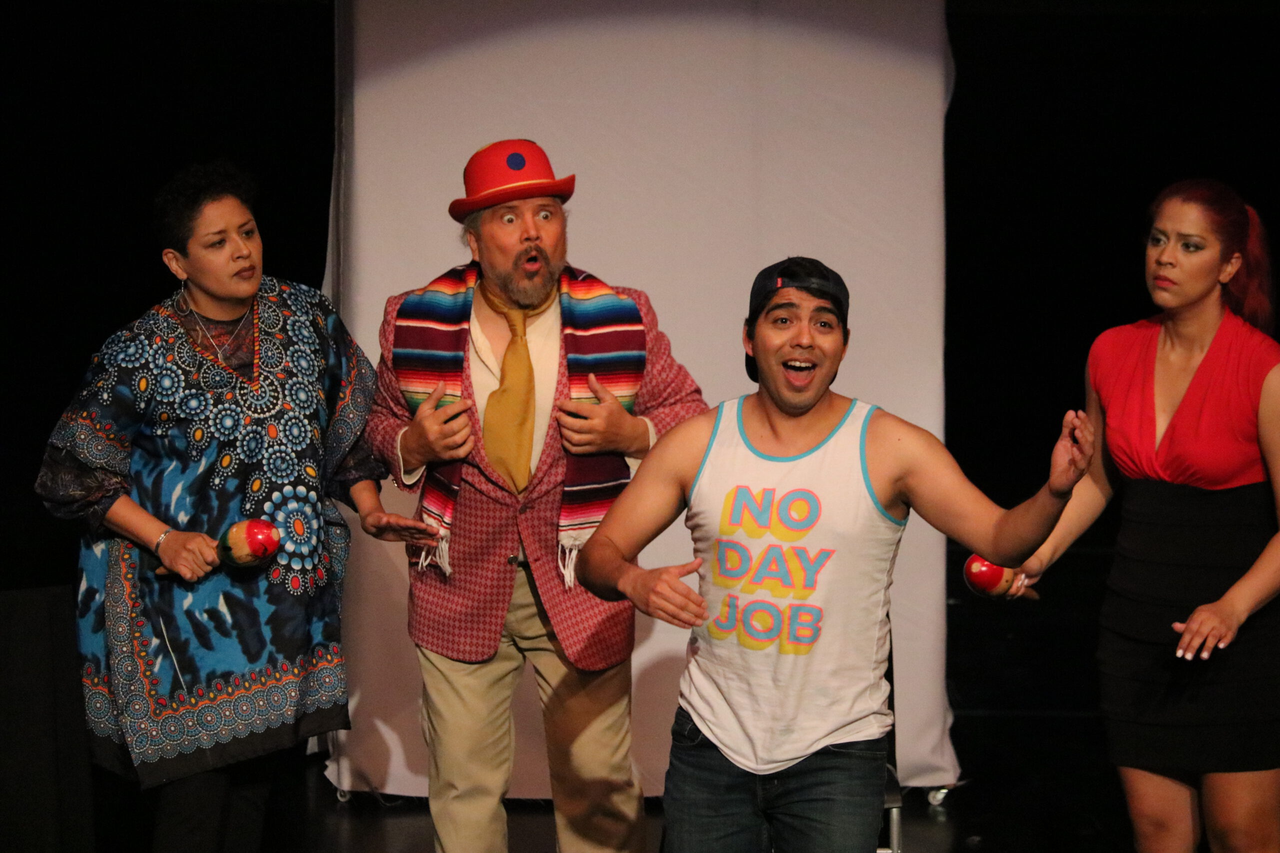 Four actors with animated expressions perform on a stage in colorful costumes. One man wears a red hat and patterned scarf, while another wears a tank top that reads, "NO DAY JOB."