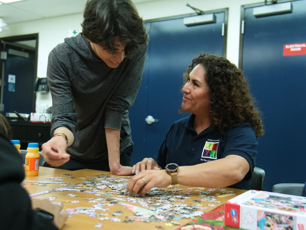 A seated woman with curly hair smiles up at a young person who is standing and leaning over a table, as they work together on a jigsaw puzzle.