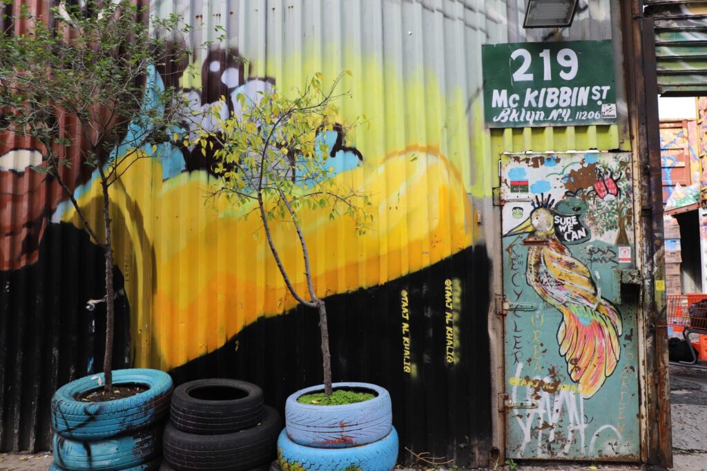 The graffiti-covered entrance to Sure We Can at 219 McKibbin Street in Brooklyn. Young trees grow from planters made of stacked, painted tires in front of a colorful mural on a corrugated metal wall.